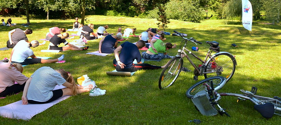 Grupa osób siedzi na trawie w pozycjach jogi. Wokół drzewa.
